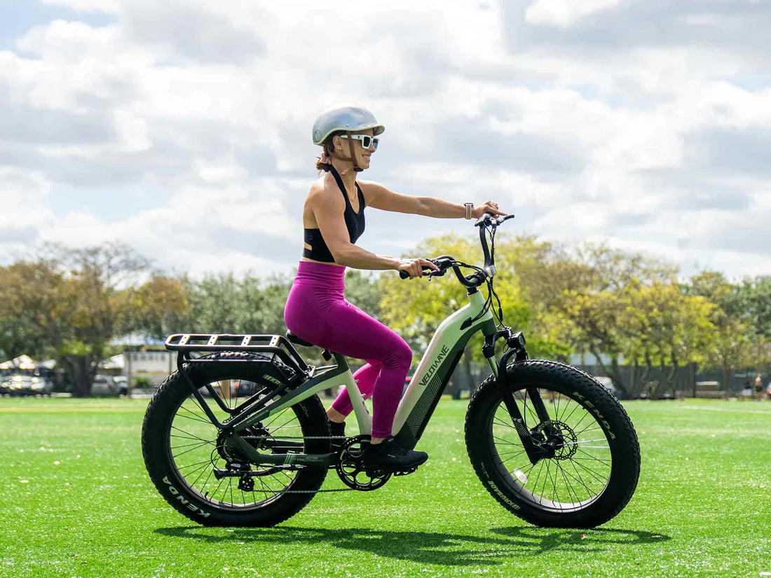 Velowave Grace 2.0 Step-Thru Electric Bike Woman riding an electric bike on a grassy field with trees and buildings in the background. Valowave Grace 2.0 Step-Thru Electric Bike