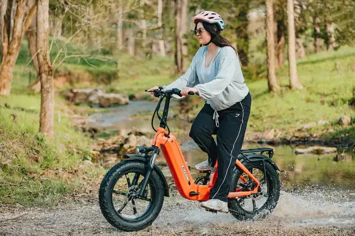 YOUNG Electric E-Flow 750W Folding eBike Person riding an orange electric bike through a forested area with a stream. YOUNG Electric E-Flow 750W Folding eBike