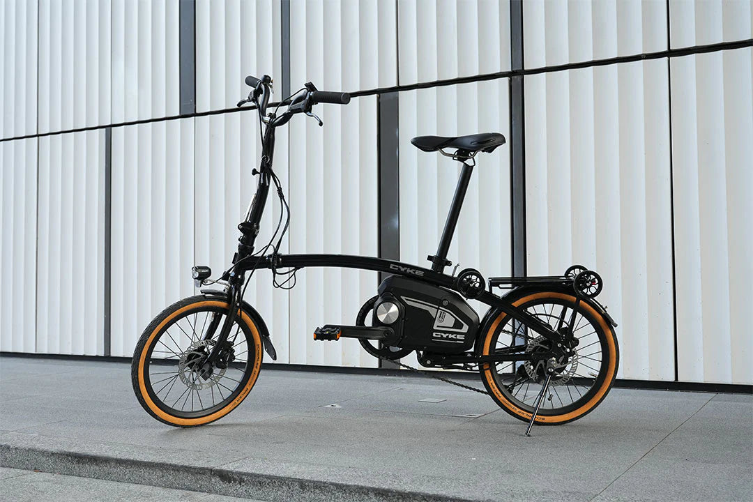 CYKE Kingfisher Mid-Drive Folding eBike Black electric bike with orange wheels on a concrete surface against a white wall. CYKE Kingfisher Mid-Drive Folding eBike