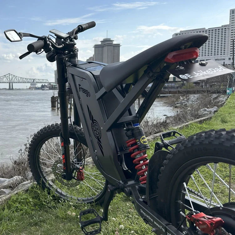 Freego X2 Pro All-Terrain Dirt eBike Electric bike with red suspension springs on a grassy area by a waterfront with buildings in the background. Freego X2 Pro All-Terrain Dirt eBike