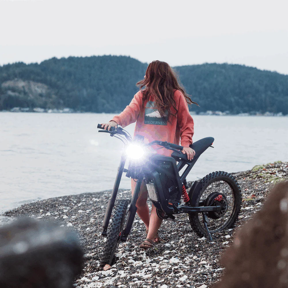 Freego X2 Dirt Master Off-Road eBike Person with a bicycle on a pebbly beach with water and trees in the background. Freego X2 Pro All-Terrain Dirt eBike