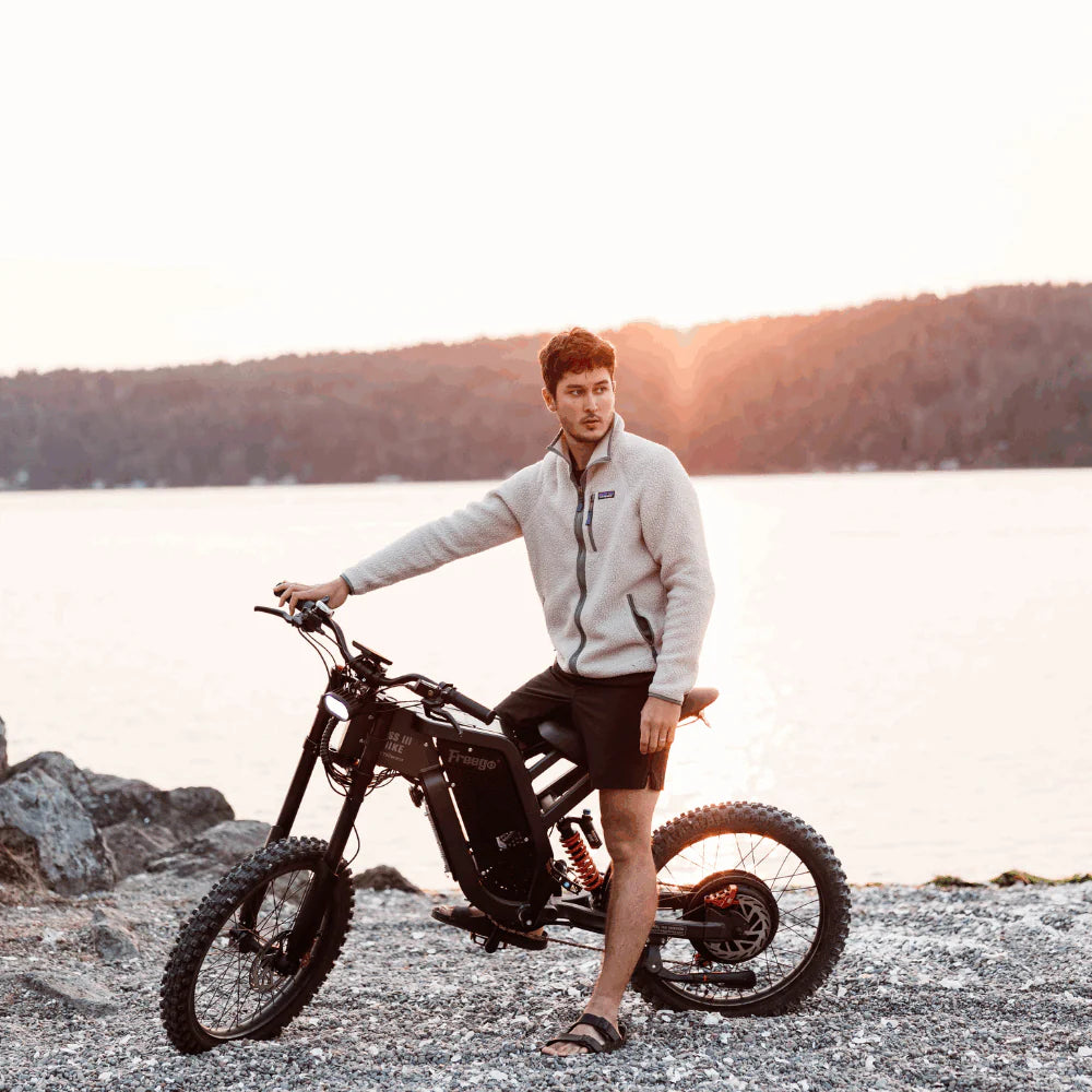 Freego X2 Dirt Master Off-Road eBike Person standing next to a motorcycle on a rocky shore with a scenic background. Freego X2 Pro All-Terrain Dirt eBike