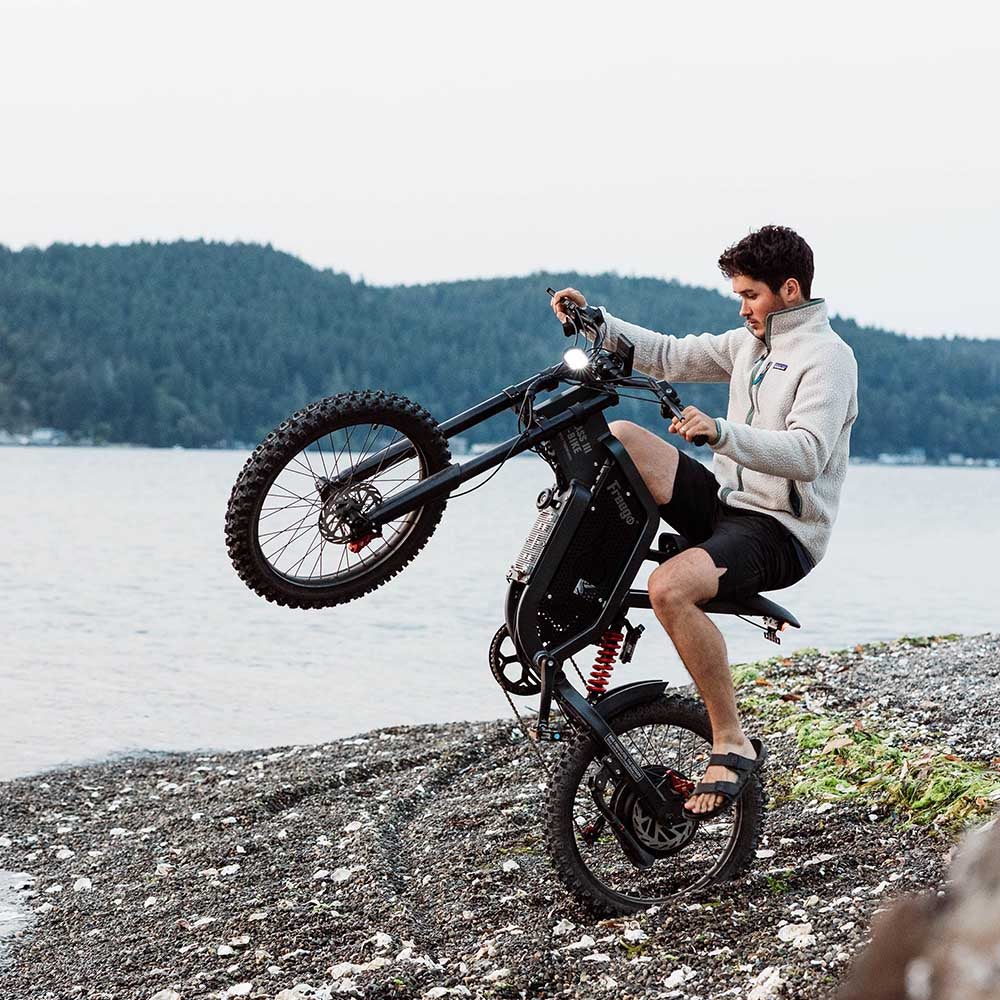 Freego X2 Dirt Master Off-Road eBike Person sitting on a unique bicycle by a lake with mountains in the background. Freego X2 Pro All-Terrain Dirt eBike