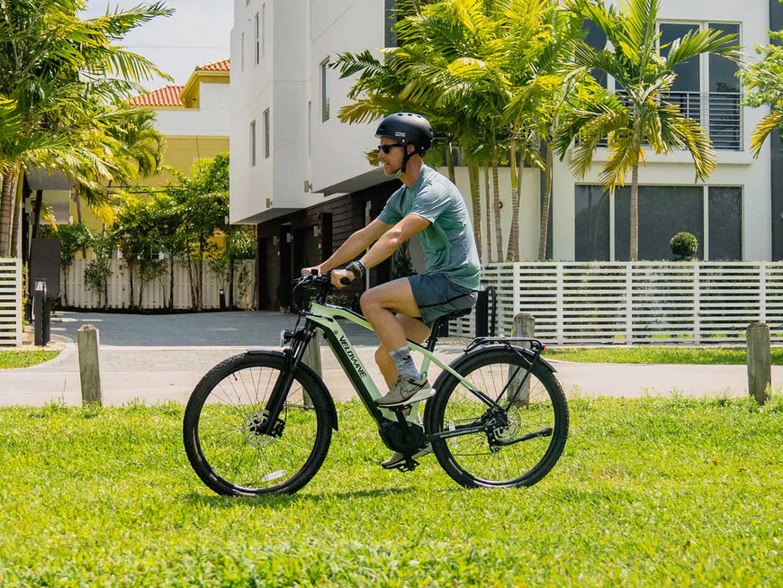 Man riding an electric bike on a grassy area with buildings and palm trees in the background. Valowave Swift M Mid-Drive Electric Bike