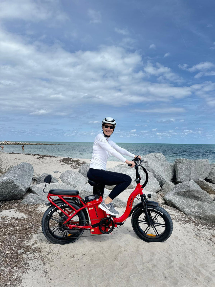 Person riding a red electric bike on a beach with ocean and sky in the background. Rattan LF Ultra Folding ebike