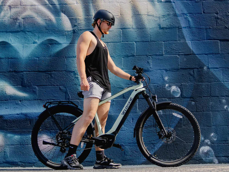 Person with a helmet standing next to an electric bike against a blue brick wall. Valowave Swift M Mid-Drive Electric Bike