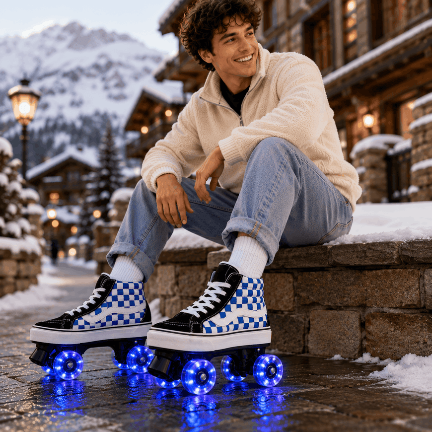 A smiling man sits on a stone ledge in a snowy mountain village, wearing black and white checkered high-top roller skates with glowing blue wheels. He is dressed in a cream fleece jacket, light blue jeans, and white socks, with snow-covered buildings and mountains in the background.