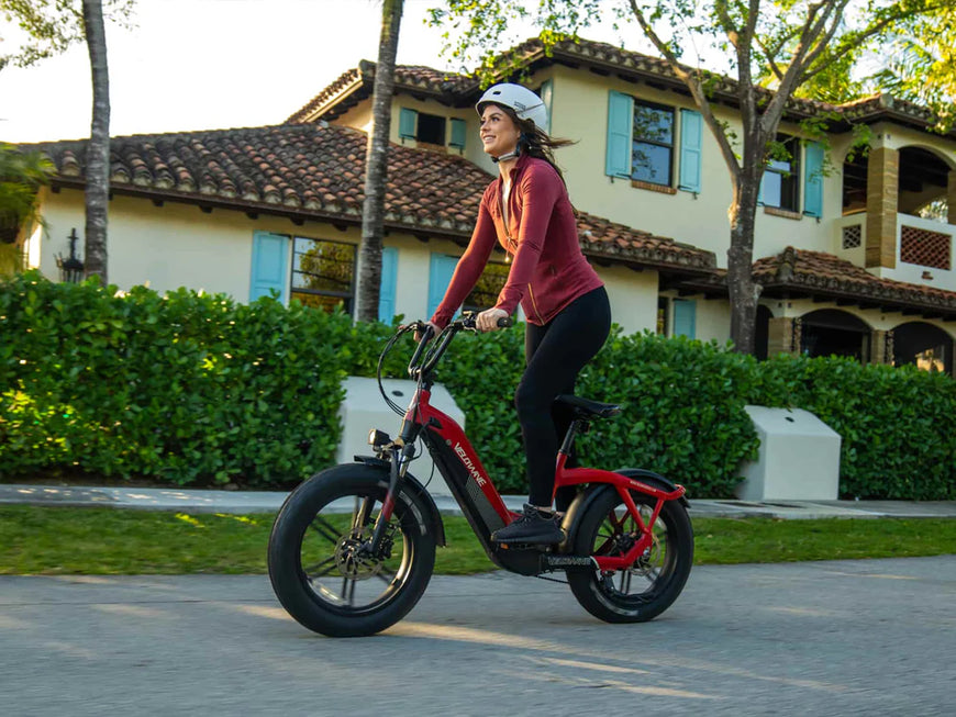 Woman riding a red electric bike on a sidewalk with a house in the background. Valowave Pony Compact Step-Thru Electric Bike