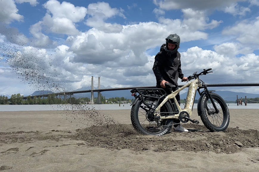 Person on an electric bike on a sandy beach with a bridge and cloudy sky in the background. CYKE Cheetah Hunting EBike