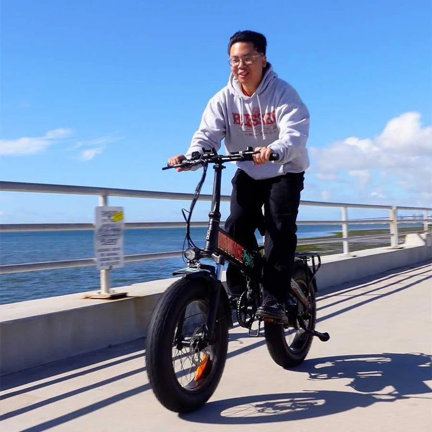 Person riding an electric bike on a bridge with a blue sky and water background. Freego eFlex Raptor E1 Shimano 7-Speed Foldable Fat Tire City Electric Bike