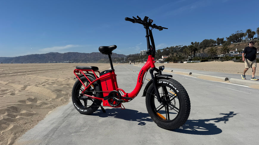 Red electric bike on a paved path with a beach and mountains in the background. Rattan LF Ultra Folding ebike