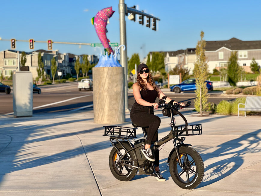 Woman riding a electric bike on a sidewalk with a large bell and colorful object in the background. Rattan LM Ultra Folding ebike