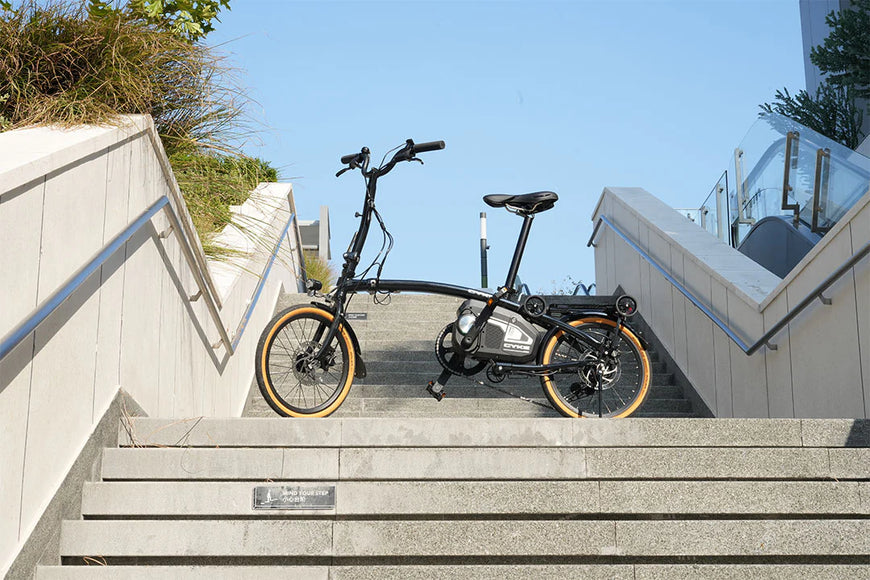 Foldable bicycle on a set of stairs with a clear blue sky in the background. CYKE Kingfisher Mid-Drive Folding eBike