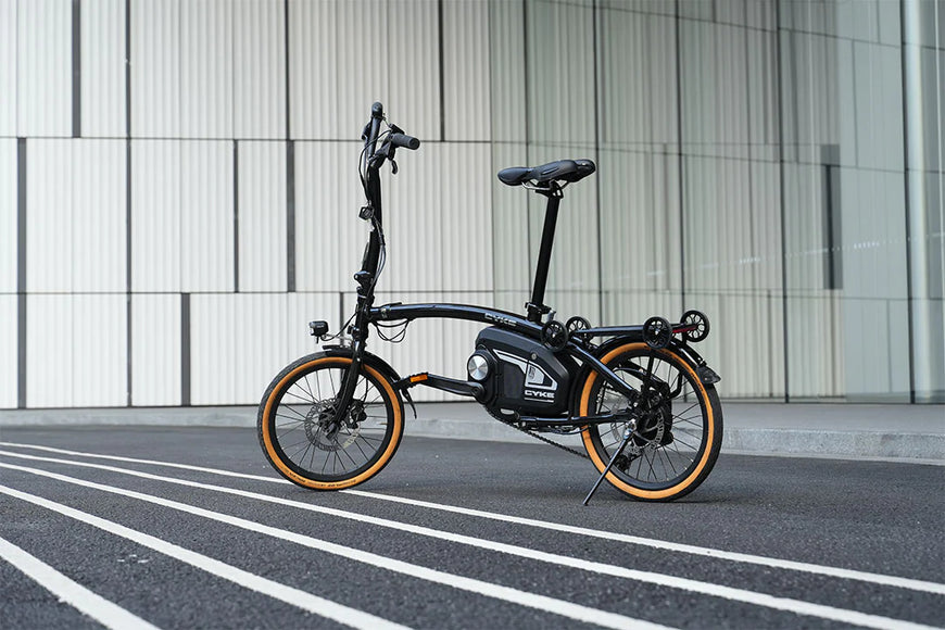 Black electric bike with orange wheels on a city street. CYKE Kingfisher Mid-Drive Folding eBike