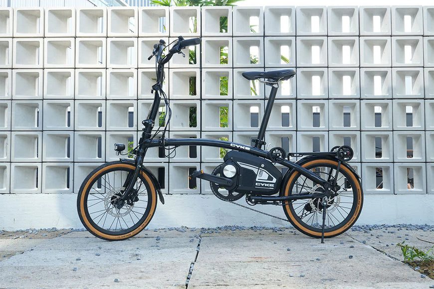 Black folding bicycle leaning against a concrete wall with square patterns. CYKE Kingfisher Mid-Drive Folding eBike