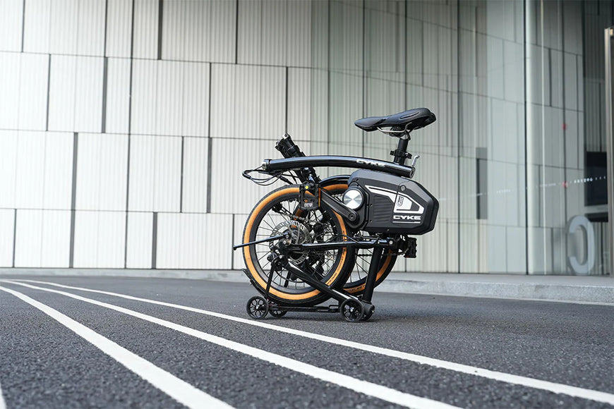 Foldable electric bike on a city street with modern building facade. CYKE Kingfisher Mid-Drive Folding eBike