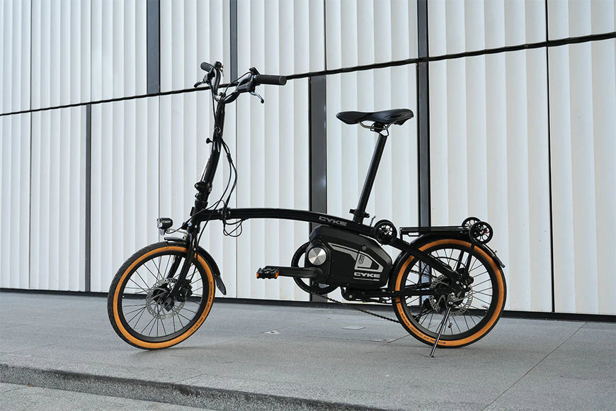 Black electric bike with orange wheels on a concrete surface against a white wall. CYKE Kingfisher Mid-Drive Folding eBike