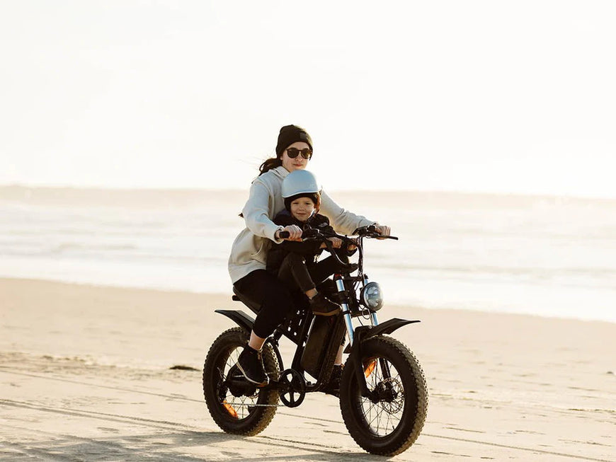 Two people riding a black electric bike on a sandy beach. Freego S1 Cafe Racer eBike
