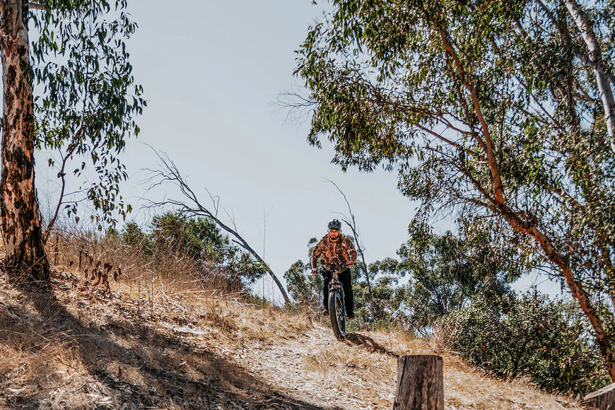 Person riding a bike on a dirt trail with trees and clear sky in the background. CYKE Cheetah Hunting EBike