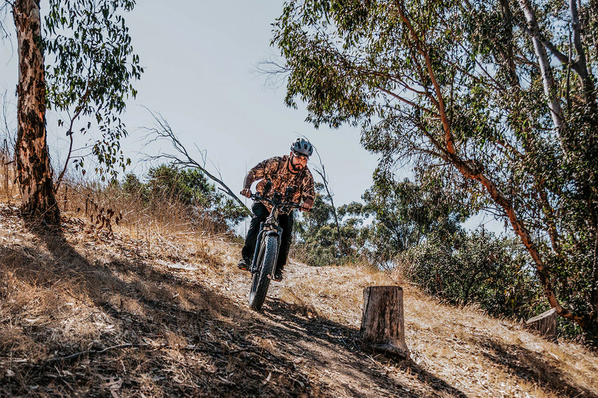 Person riding a mountain bike on a trail with trees and a clear sky. CYKE Cheetah Hunting EBike