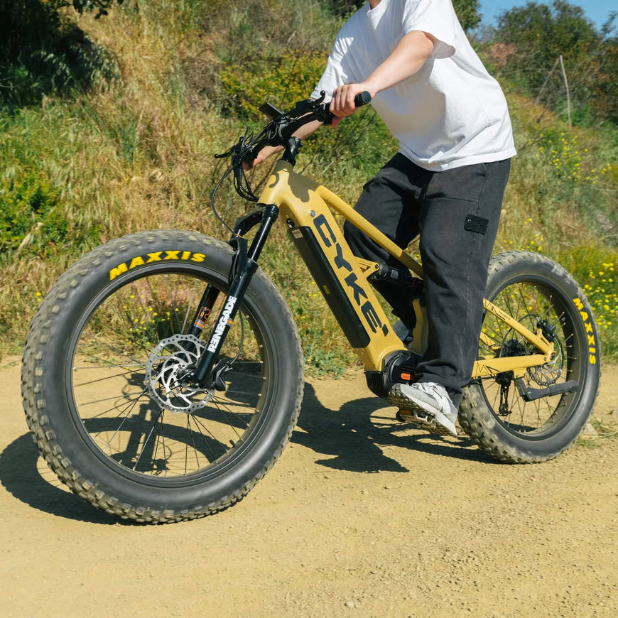 Person riding a yellow electric bike on a dirt path with greenery in the background. CYKE Falcon X Full Suspension EBike