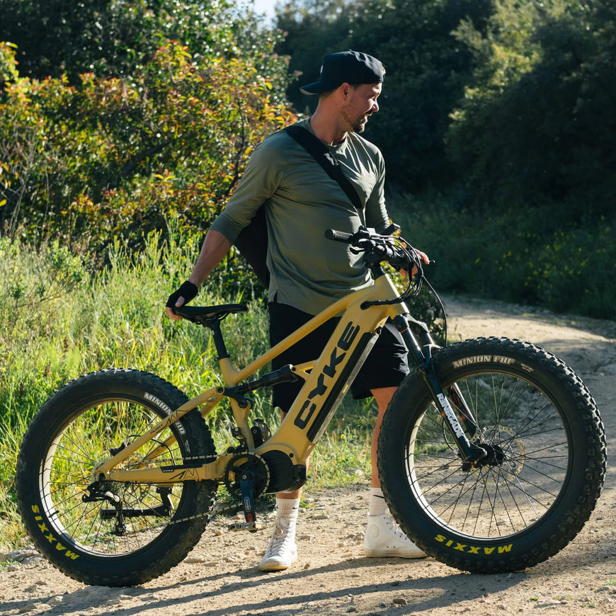 Man holding a yellow fat-tire bike on a dirt path with greenery in the background. CYKE Falcon X Full Suspension EBike