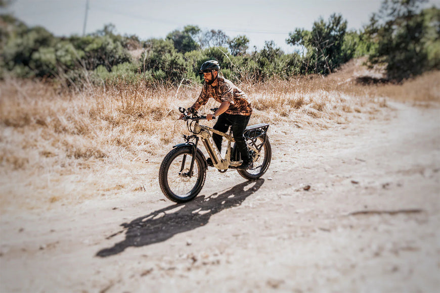 Person riding a bike on a dirt path with dry grass and trees in the background. CYKE Cheetah Hunting EBike