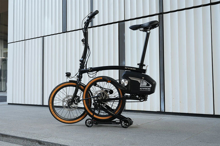Foldable electric bike on a stand against a modern building. CYKE Kingfisher Mid-Drive Folding eBike